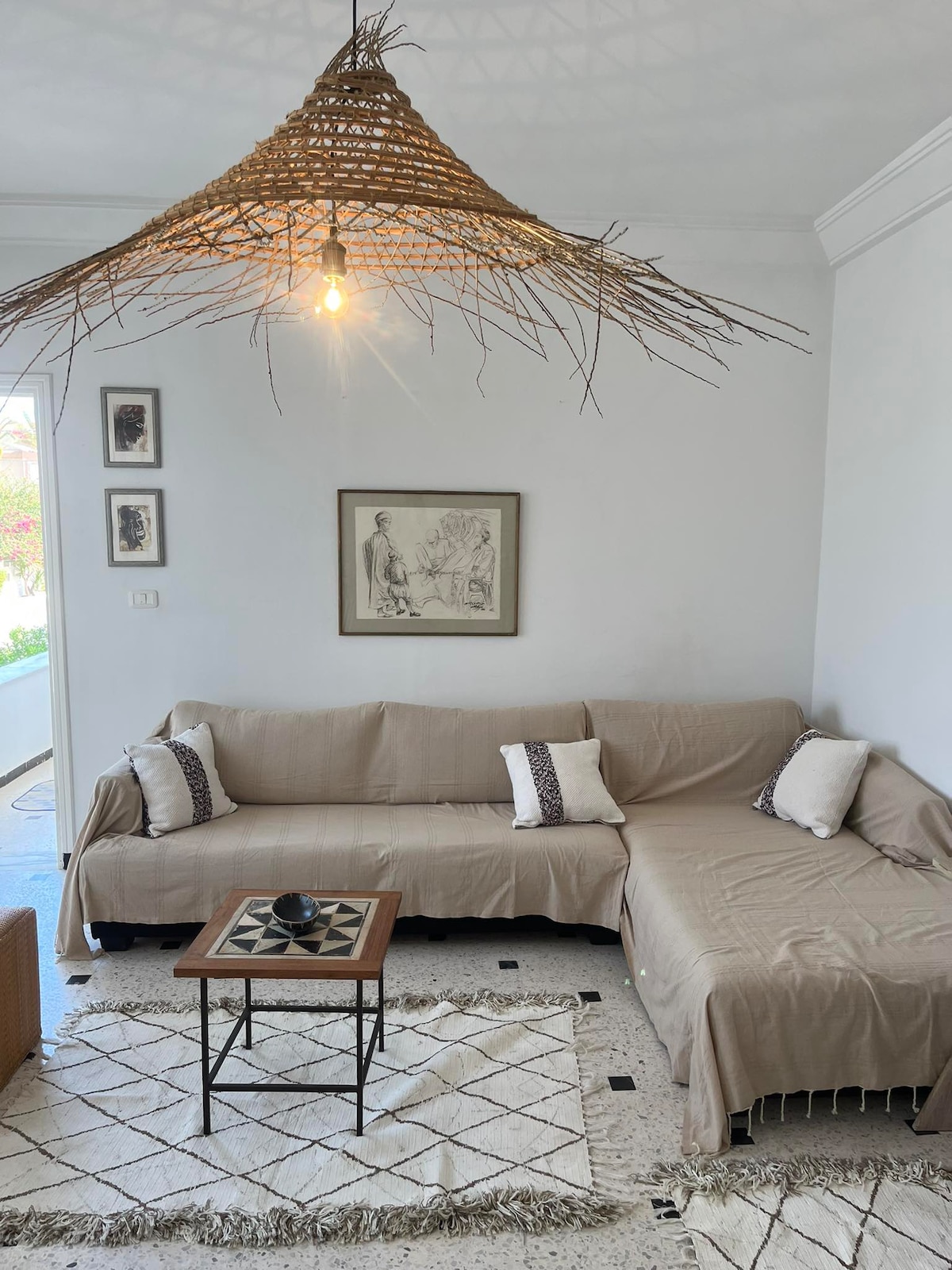 A comfortable living area features a light-colored sectional sofa adorned with patterned cushions. A wooden coffee table sits atop a textured rug, accompanied by a decorative black candle holder. Natural light enters through nearby doors, illuminating the space's simple and serene decor.