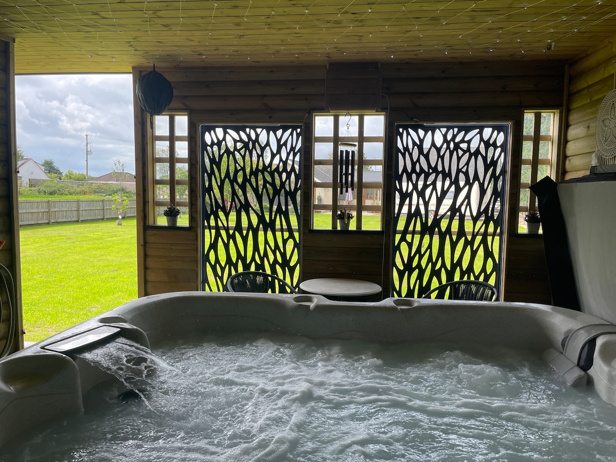 A hot tub is visibly set within a wooden structure, featuring intricate partition screens that provide a stylish backdrop. Lush green grass is seen outside through open space, with a table and chairs for relaxed seating nearby.