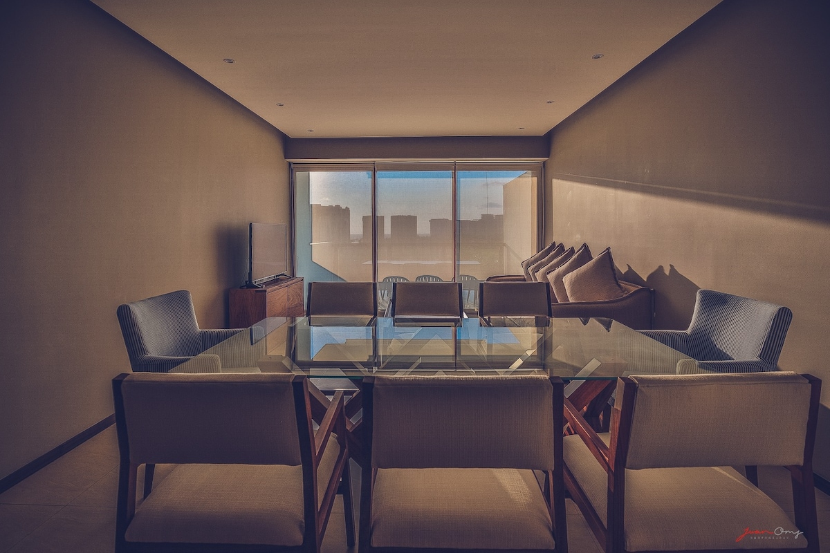 A spacious living area features a glass-top dining table surrounded by wooden chairs. Soft seating, including large cushions, is positioned against the large windows, which provide a view of the outdoors. A flat-screen TV is mounted on a wooden cabinet along the wall.