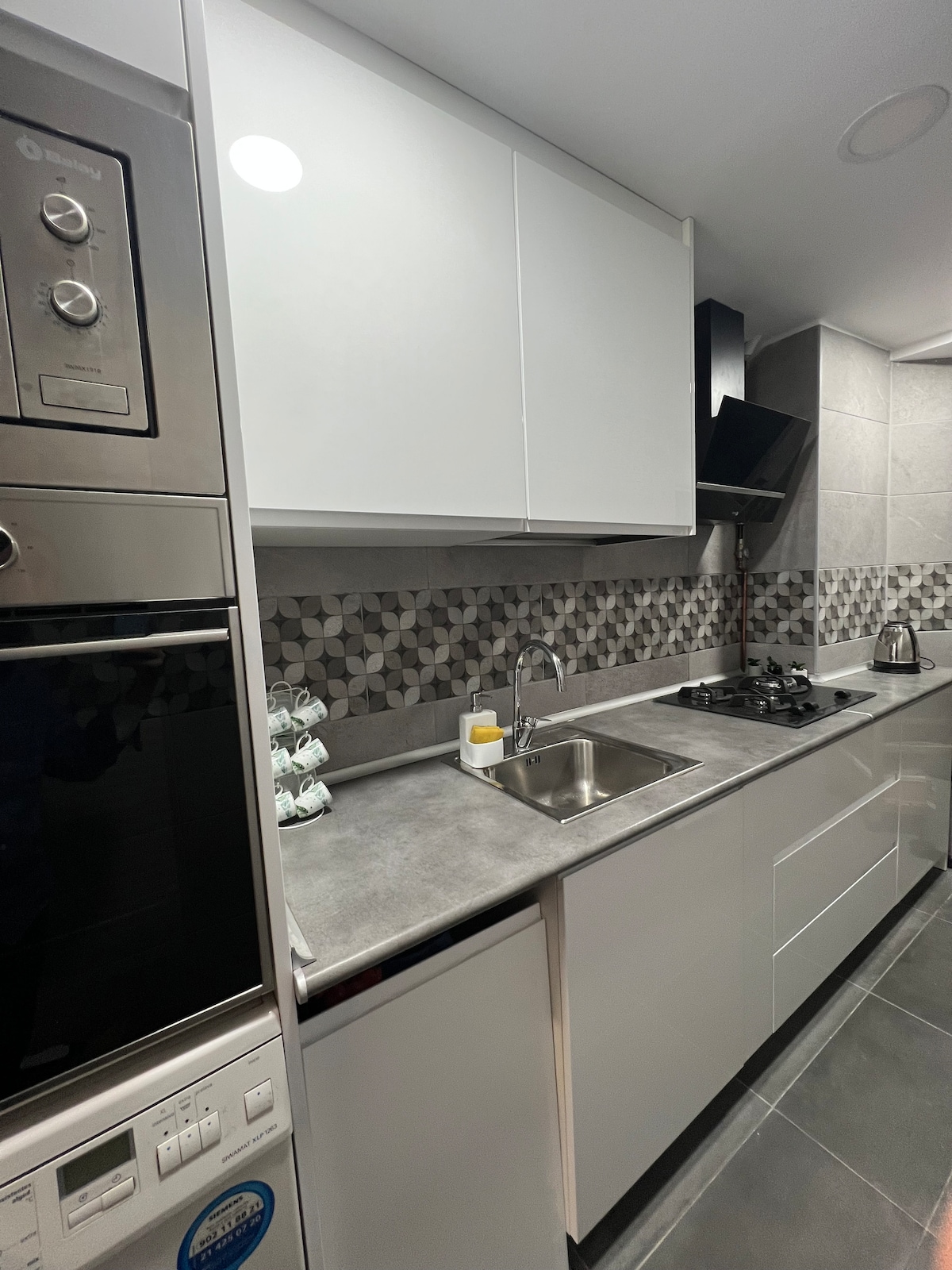 A modern kitchen is displayed with sleek white cabinetry and a grey countertop. An oven, microwave, and gas stove are integrated into the design. A small sink is situated beneath wall-mounted cabinets, and patterned backsplash tiles add visual interest.