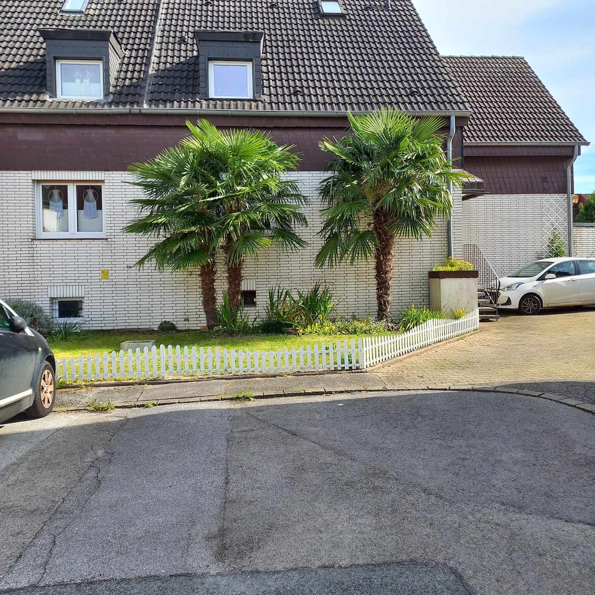 The exterior displays a modest garden with two palm trees framed by a low white picket fence. A light-colored building is visible in the background, with multiple windows featuring white curtains. A small parking area accommodates vehicles nearby.