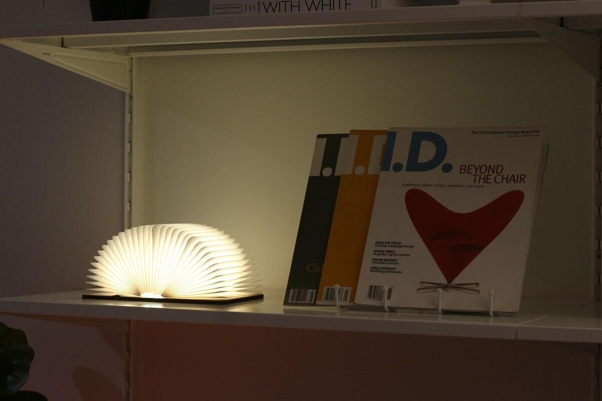 A stylish lamp emits a soft glow, creating visual interest on a shelf. Nearby, several magazines are neatly arranged, their colorful covers contrasting with the lamp's warm light. The overall composition highlights a minimalist and modern design aesthetic.