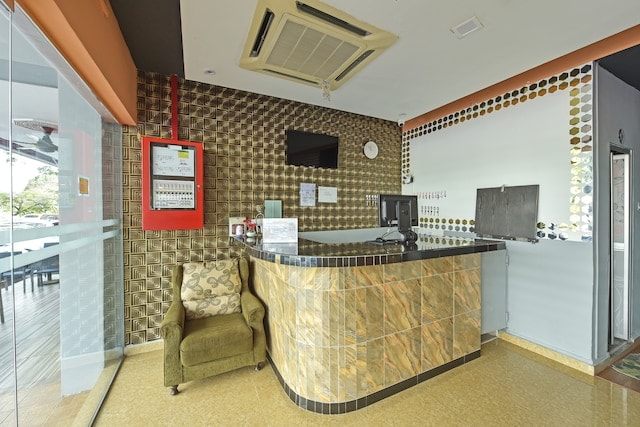 A reception area is shown with a curved counter featuring a patterned finish. A green chair is positioned beside the counter. The walls are adorned with a mix of metallic tiles and a bulletin board is affixed near the entrance, adding a functional element to the space.