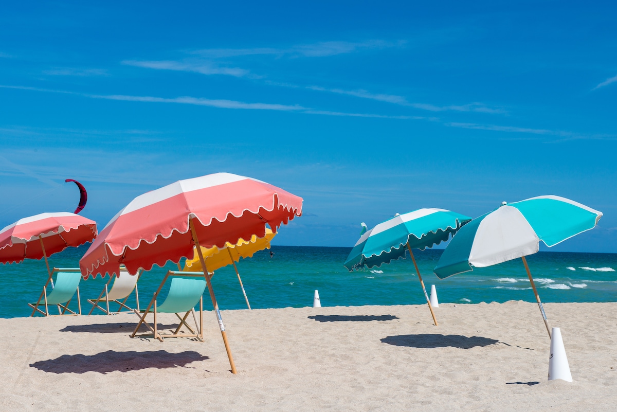 Colorful beach umbrellas are arranged in a serene sandy area, contrasting with the clear blue ocean in the background. Lounge chairs are placed beneath the umbrellas, inviting relaxation. The gentle waves and clear sky enhance the tranquil beach setting.