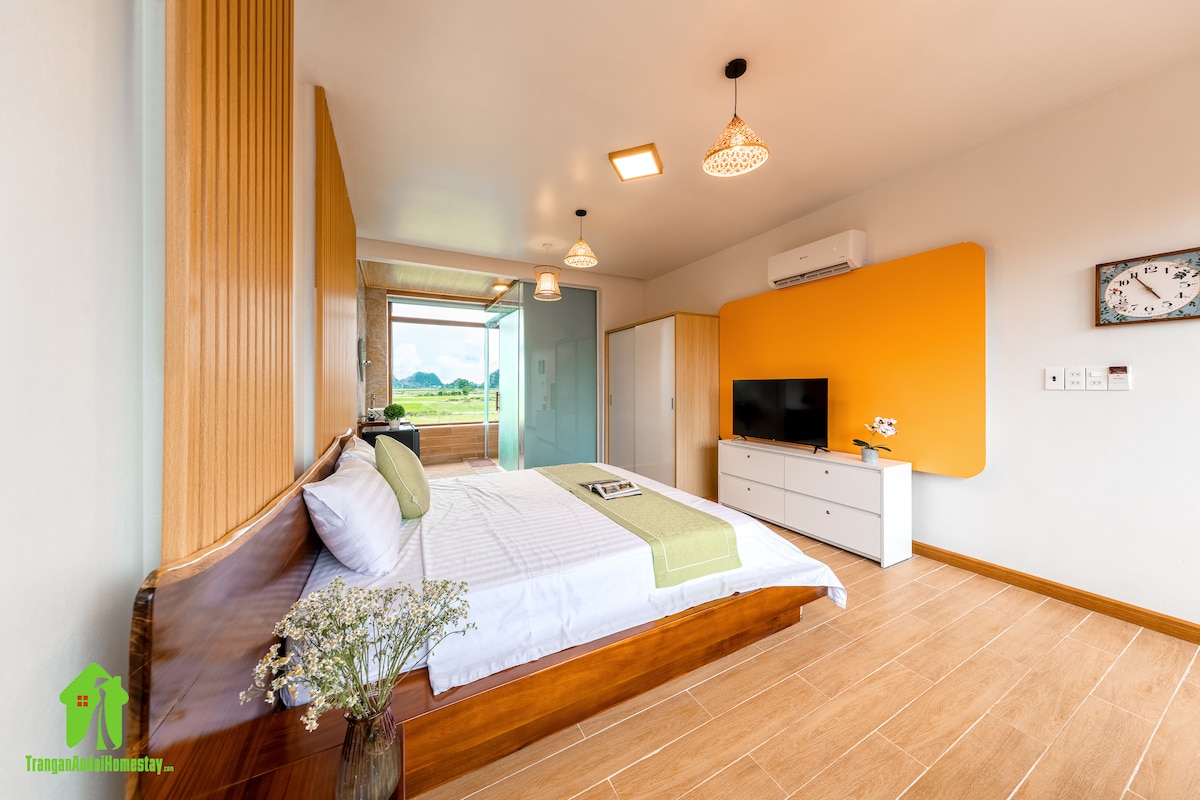 A spacious sleeping area features natural wood accents and a large bed with crisp linens. A vibrant orange wall enhances the room, while a wall-mounted TV and minimalist furniture provide functionality. Large glass doors offer views of the surrounding mountains.
