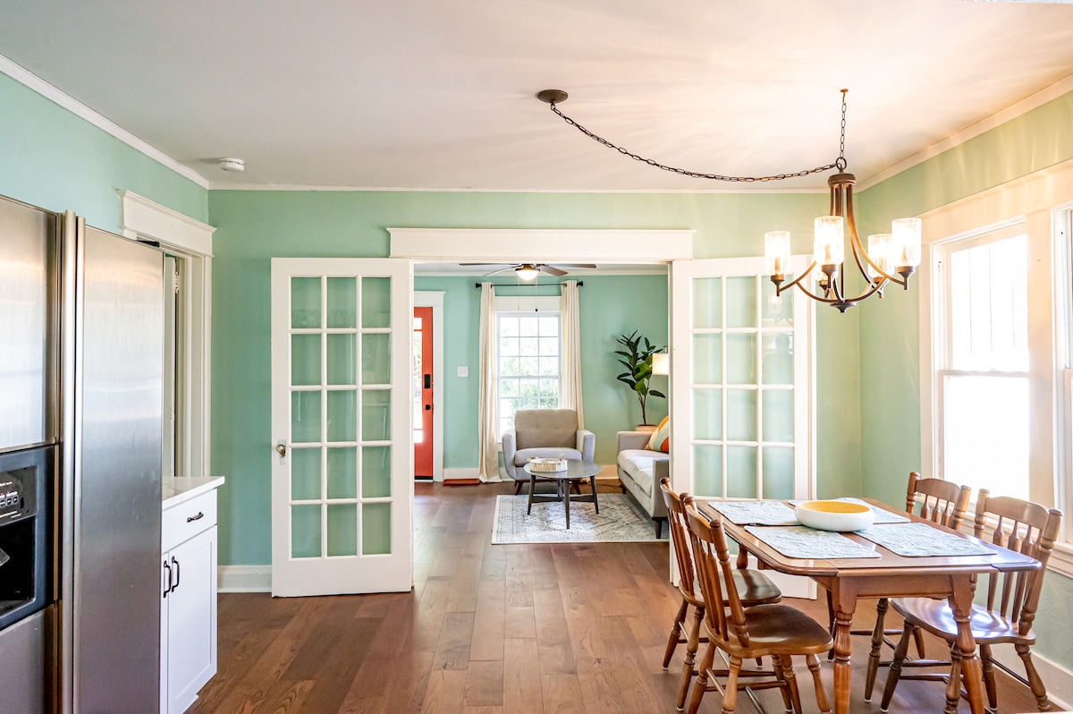 A bright dining area features a wooden table with seating for six. French doors separate it from the living space, showcasing a cozy seating arrangement and natural light. Soft green walls and hardwood flooring create a welcoming environment.