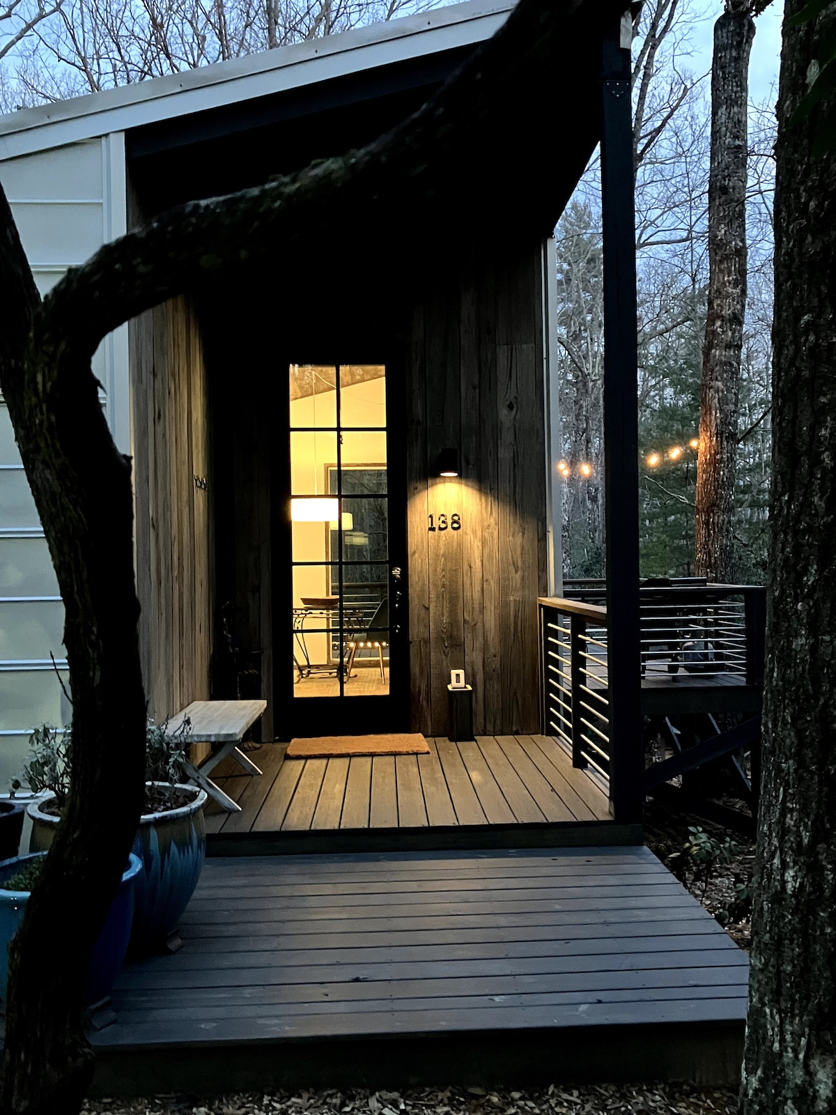 A welcoming entrance displays a simple wooden deck leading to a door marked '138', illuminated by a soft light. Lush greenery and potted plants frame the pathway, while gentle evening light highlights the elements of the cottage nestled among the trees.