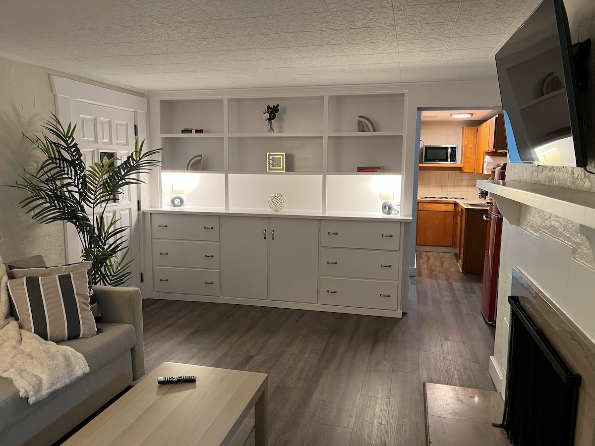 The living area features a gray sofa with light throw blankets and a small coffee table. Shelving units with decorative items and drawers are present in the background. A glimpse of a well-equipped kitchen is visible through an archway, enhancing the home-like environment.