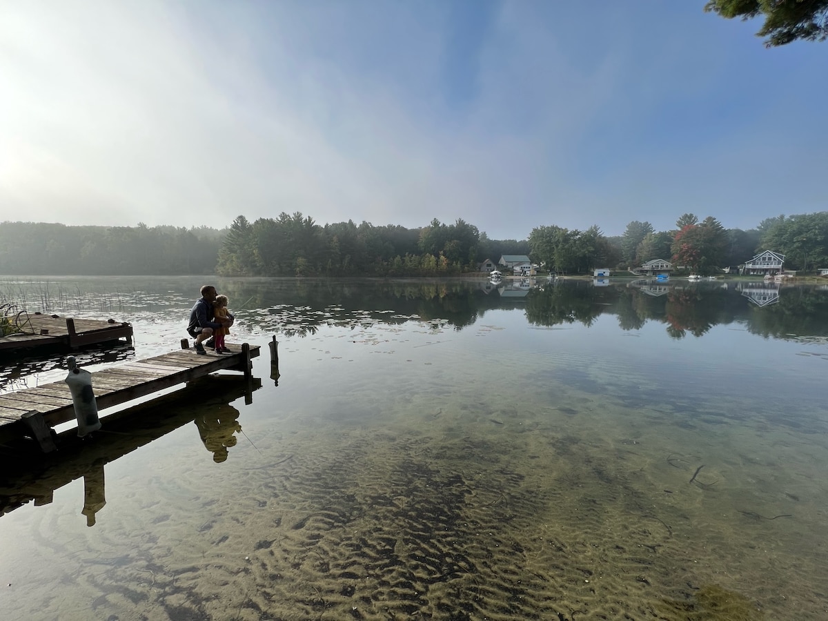 A wooden dock extends over calm waters, reflecting the surrounding trees and houses. Two individuals sit at the edge, observing the tranquil lake. Morning mist hovers above the water, creating a serene atmosphere.