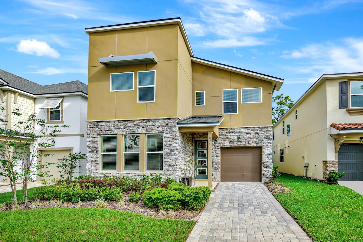 A modern two-story home features a blend of cream-colored stucco and stone accents. Large rectangular windows adorn the front, allowing natural light to enter. A paved pathway leads to the front door, flanked by neatly trimmed greenery and shrubs, contributing to the inviting curb appeal.