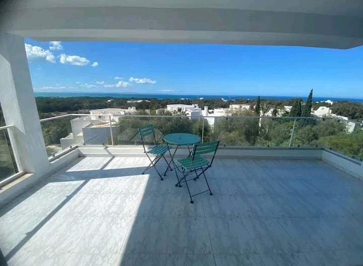A sunlit terrace features a small round table surrounded by two green chairs, offering views of the surrounding landscape and distant ocean. The area is paved with light-colored tiles, and the glass railing enhances the open feel.