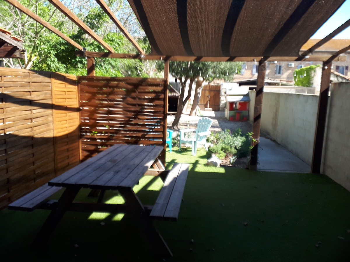 An outdoor space features shaded seating with a wooden picnic table and white adirondack chairs. Lush greenery surrounds the area, with a small garden visible and a play structure in the background. A fence provides privacy, contributing to a tranquil environment.