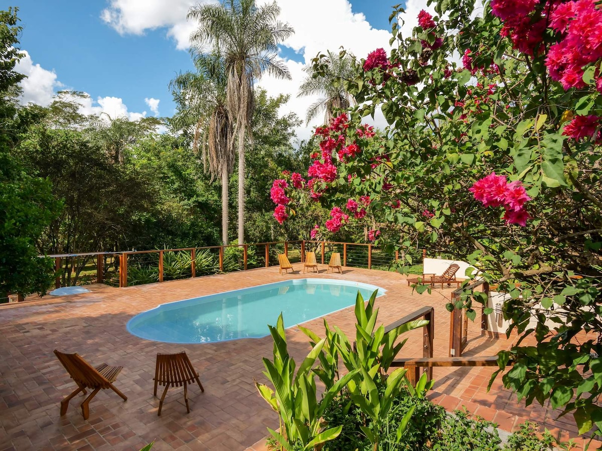 A serene outdoor space is presented, featuring a swimming pool surrounded by vibrant floral landscaping. Wooden chairs are arranged nearby, complementing the natural scenery of palm trees and lush greenery under a clear blue sky.