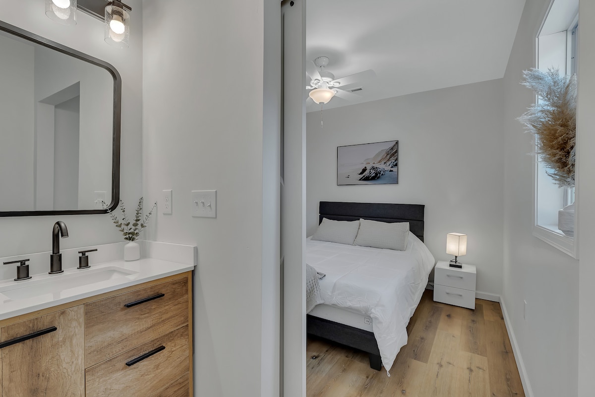 A bright bedroom is visible, featuring a spacious bed with a light-colored bedding set. A nightstand stands beside the bed, topped with a lamp. The adjoining bathroom includes a modern vanity with dual sinks and a large mirror, complemented by natural wood accents.
