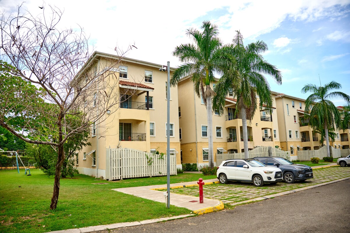 The exterior of a three-story apartment building is shown, surrounded by lush greenery. Palm trees are visible in the foreground, and a grassy area features a swing set. Vehicles are parked along a paved path, with a fire hydrant positioned nearby.