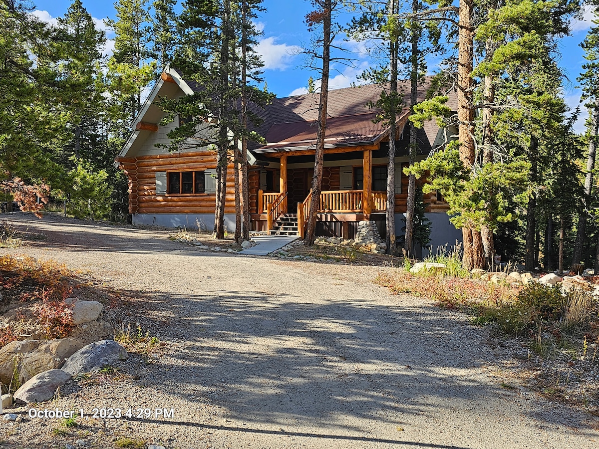 A custom-built log cabin, crafted from hand-hewn Douglas fir logs, stands amidst tall pine trees. The spacious front porch features wooden railings and steps leading to the entrance, while a gravel driveway offers easy access. Surrounding natural beauty creates a serene, secluded environment.