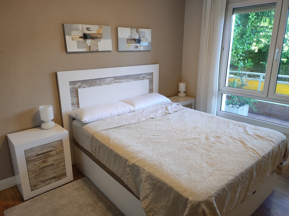 A well-appointed bedroom features a spacious bed with a light-colored quilt. Two bedside tables flank the bed, each topped with a lamp. A large window allows natural light to enhance the room, overlooking a private balcony with greenery.
