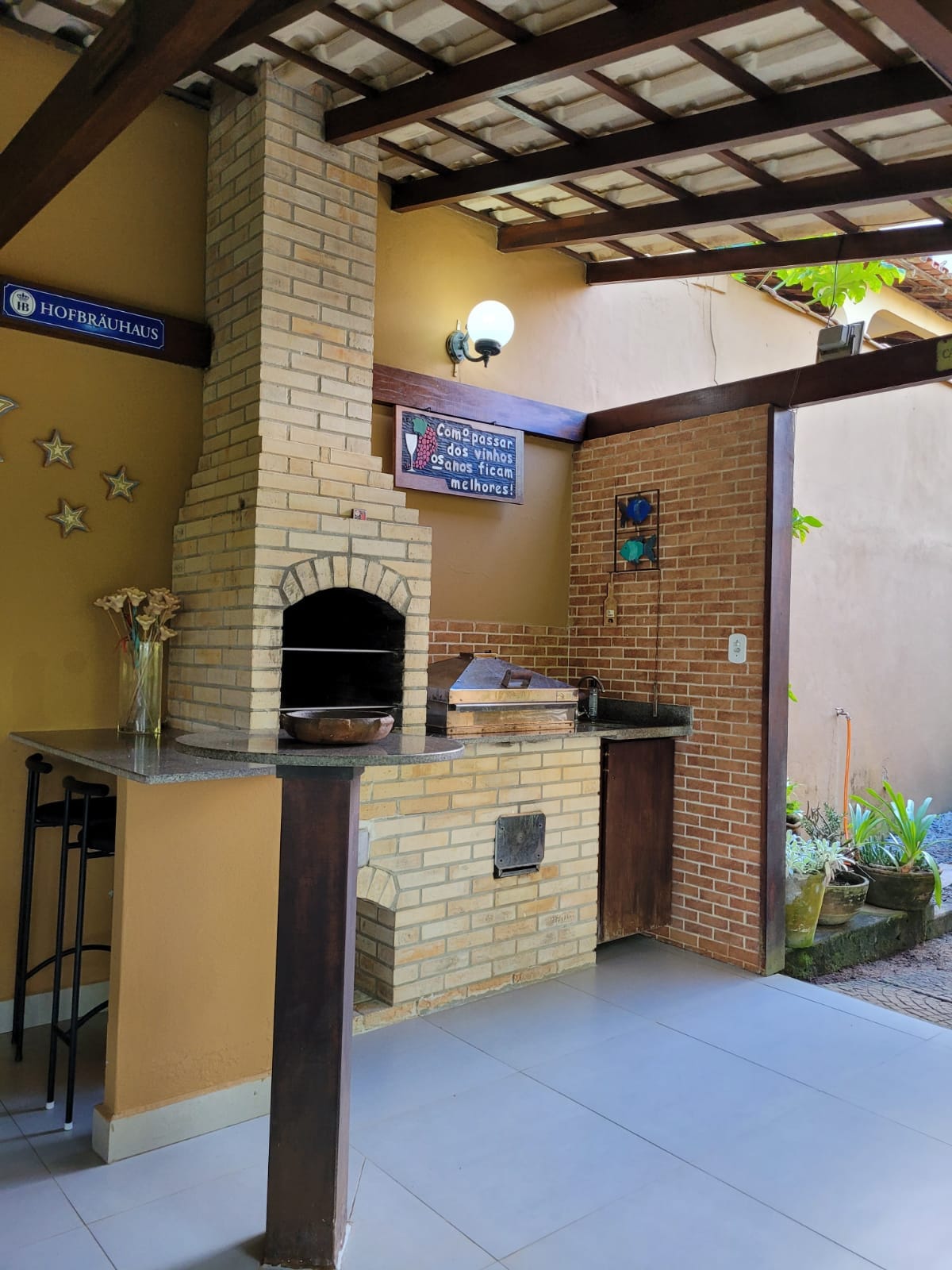 An outdoor kitchen area features a brick barbecue set against a warm-toned wall. A countertop provides space for food preparation, and decorative elements include a wall-mounted artwork. Natural light enhances the inviting atmosphere of this space, surrounded by greenery visible in the background.
