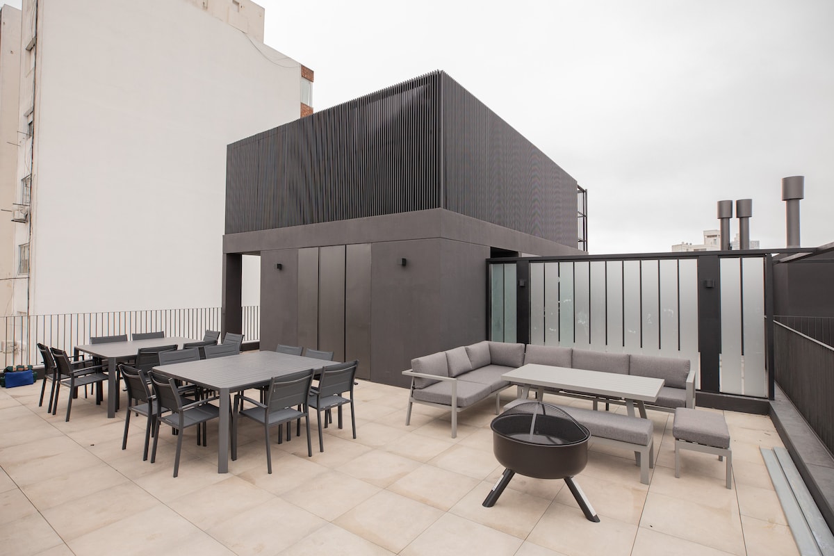 A spacious public terrace is featured, furnished with multiple seating areas and dining tables. Gray lounge chairs and sofas complement the modern aesthetic. A circular fire pit is positioned at one end, providing a cozy gathering spot. The backdrop showcases contemporary building designs.