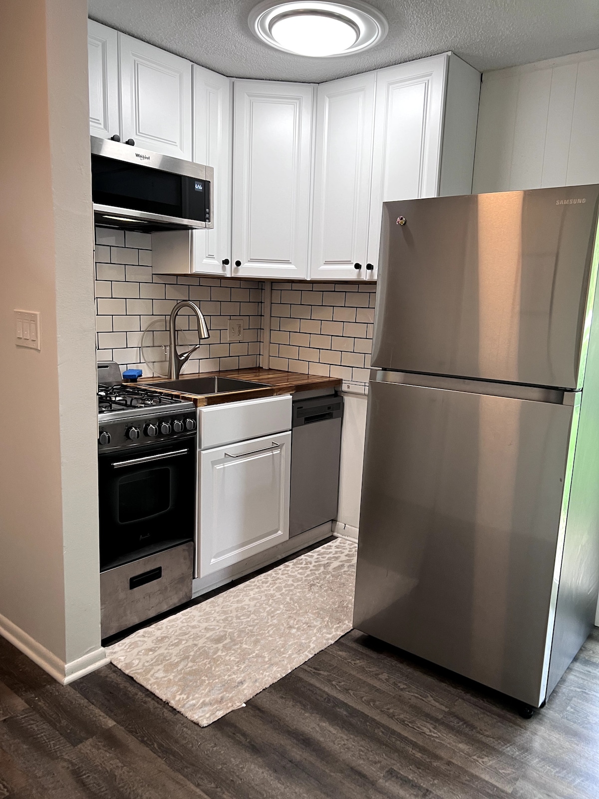 A modern kitchen area showcases sleek white cabinetry paired with a stainless-steel refrigerator and dishwasher. A gas stove is positioned beneath a microwave, with a tiled backsplash enhancing the clean lines. Soft light is provided by a ceiling fixture.