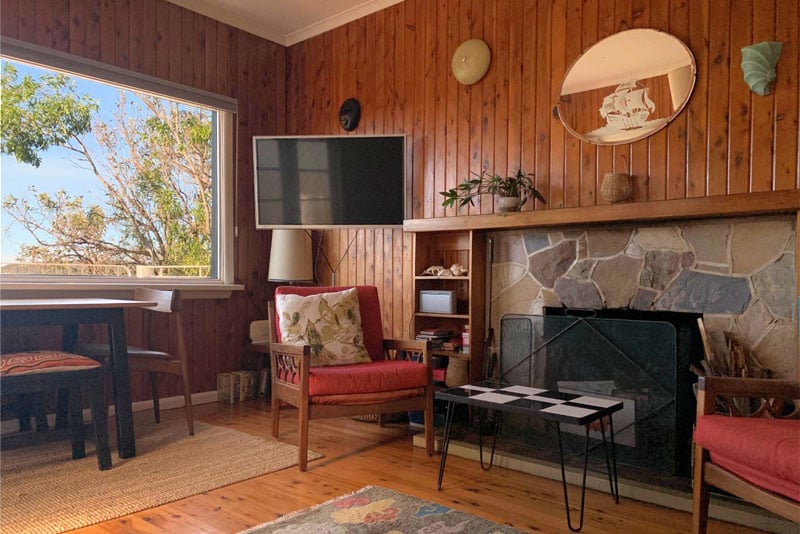 A cozy lounge area features wooden paneling and natural light from a large window. A flat-screen television is mounted on the wall above a stone fireplace. Two armchairs with colorful upholstery are positioned near a small table, while a dining area is visible in the background.