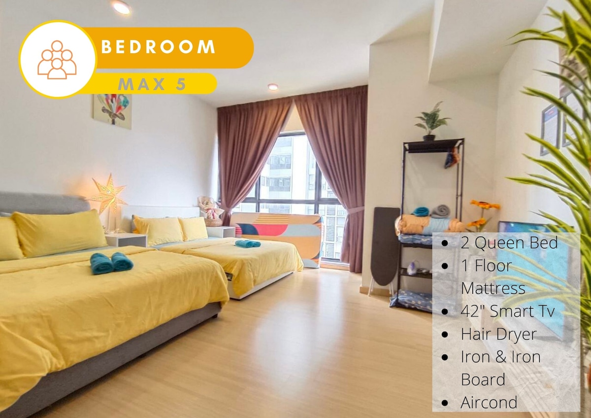 The spacious bedroom features two queen beds with yellow linens, a floor mattress, and a 42-inch Smart TV. A large window allows natural light to illuminate the room, while decorative plants and artwork add a touch of character. Air conditioning is available for comfort.