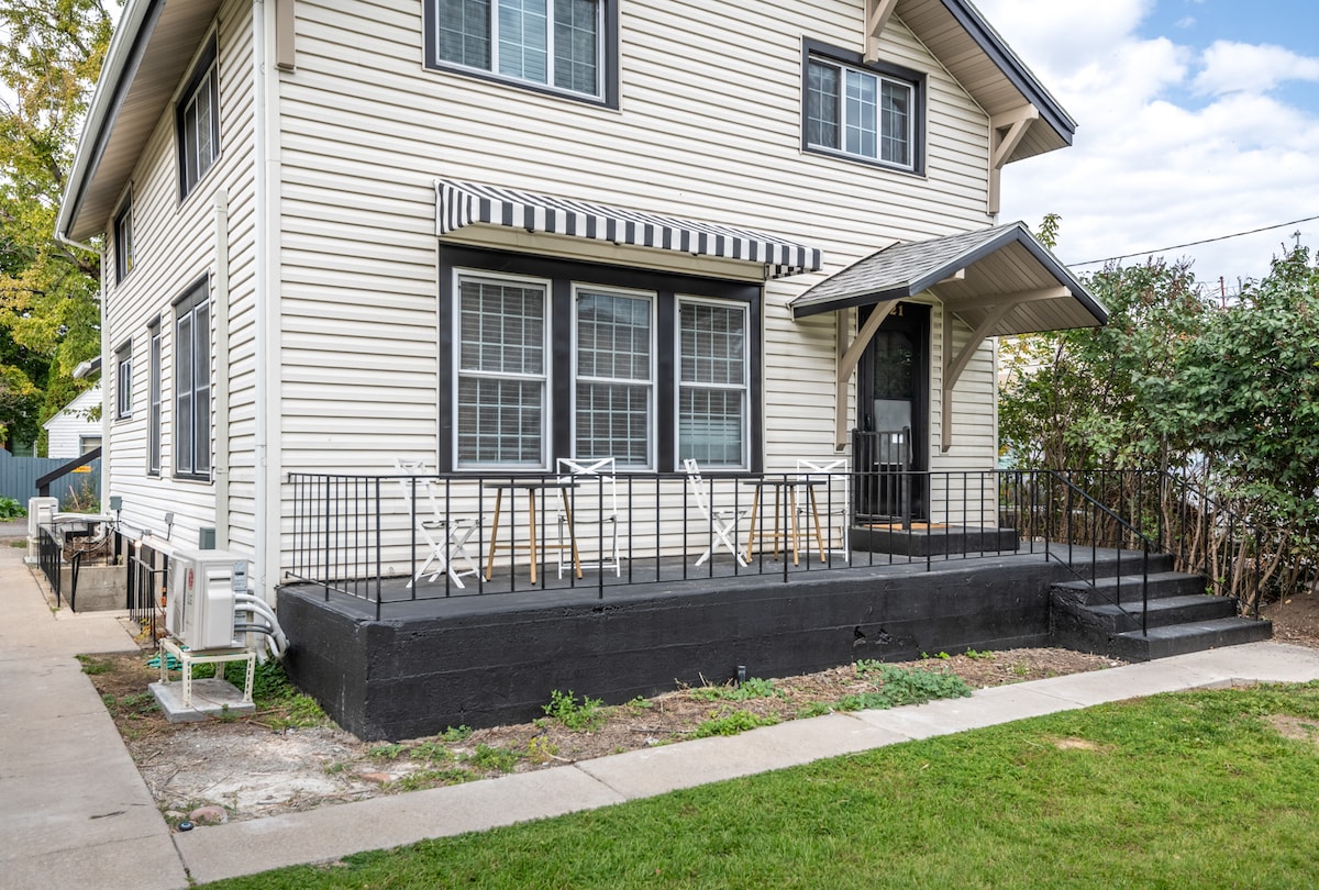 The exterior of the vintage studio apartment features a large deck with a black railing. Two white bistro chairs are positioned on the deck, accompanied by small tables. The entrance is framed by a gabled roof, with several windows adorned by striped awnings.