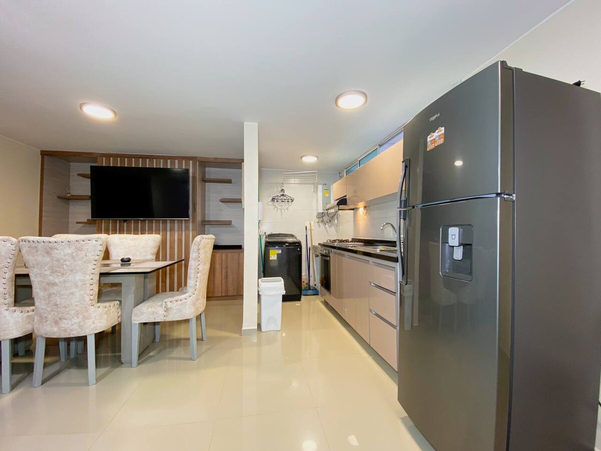 A modern kitchen area features sleek cabinetry, a large refrigerator, and a convenient dining table surrounded by plush chairs. Light-colored tile flooring enhances the spacious feel, while a television is mounted on the wall opposite the dining area.