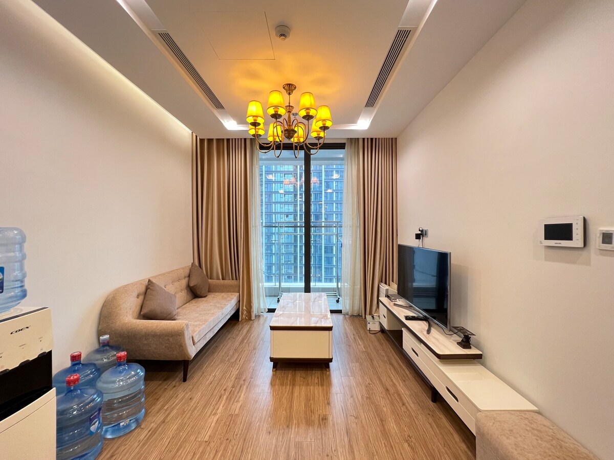 A bright living area is featured, showcasing a beige sofa and a wooden coffee table. Large windows offer city skyline views, while warm lighting is provided by a chandelier. A flat-screen TV is positioned on a white console, with water bottles arranged neatly nearby.