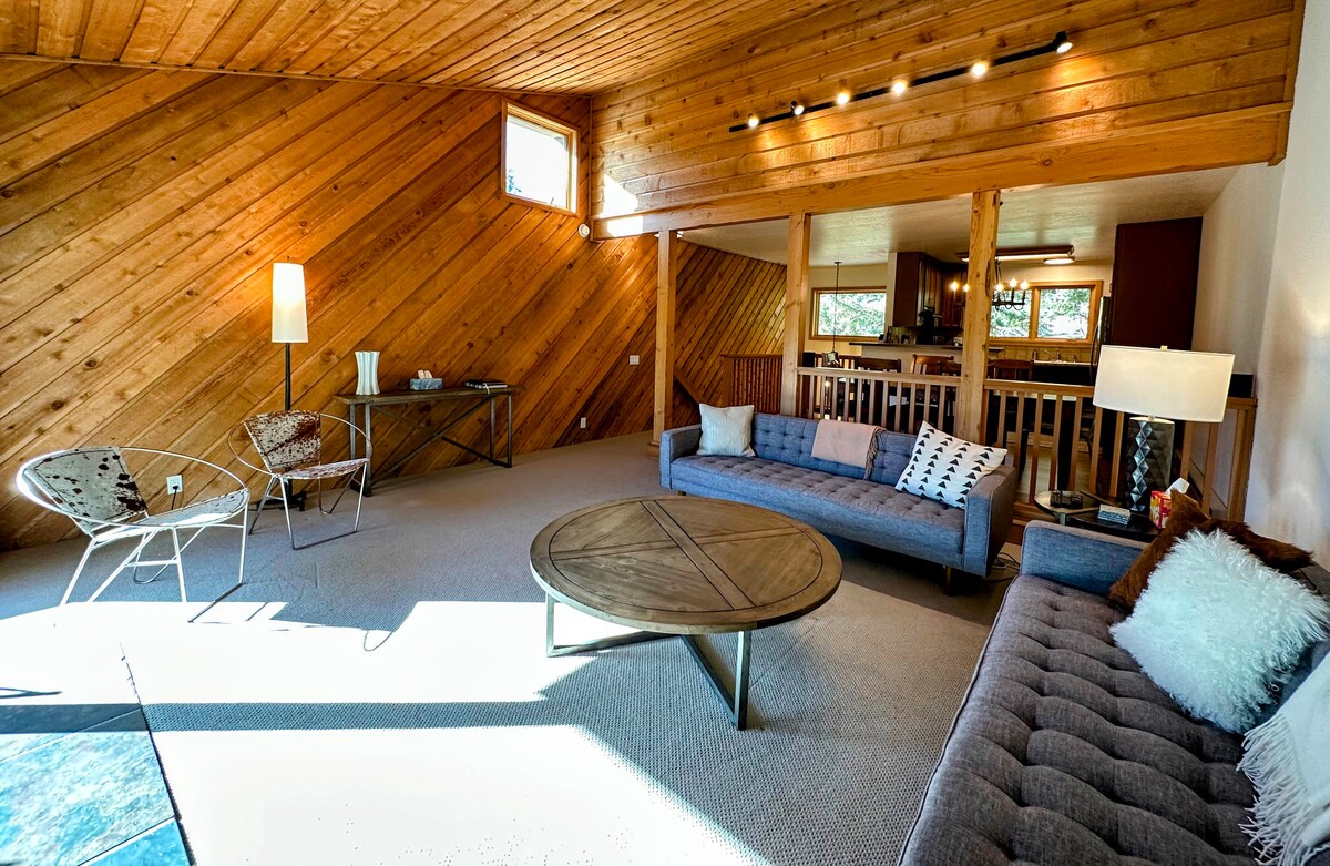 The spacious living area features a high wood-paneled ceiling that creates an open atmosphere. A plush gray sofa and a modern chair are positioned around a round coffee table, illuminated by natural light streaming in from large windows.