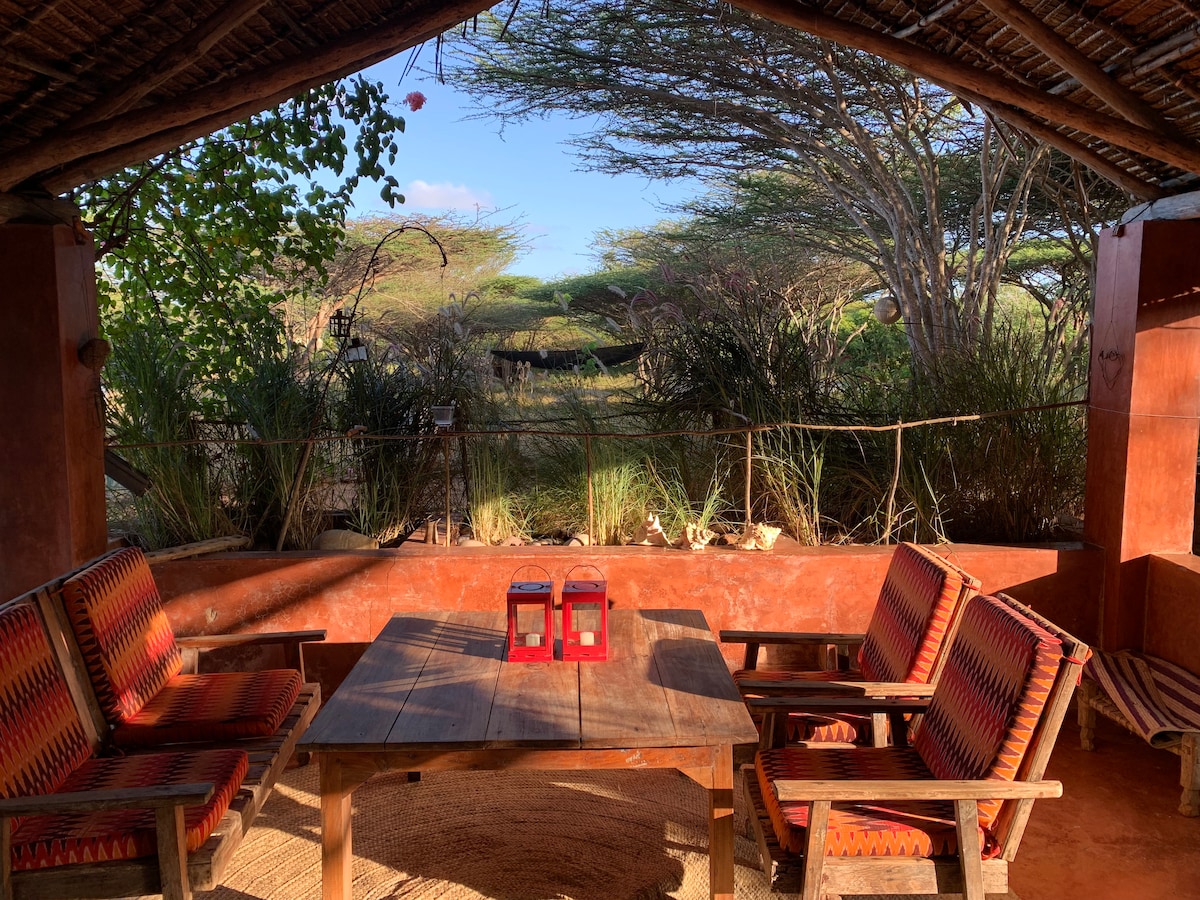 An outdoor seating area features a wooden table surrounded by chairs with traditional patterns. Two red lanterns sit atop the table. Lush greenery and open sky are visible in the background, creating a serene atmosphere.