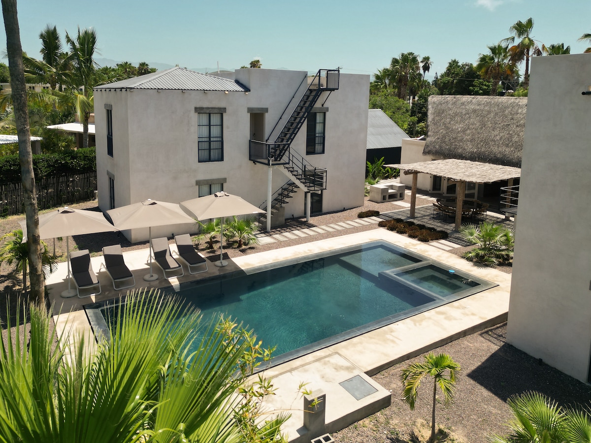 A spacious outdoor area features a custom hydrazzo pool surrounded by six loungers and landscaped gardens. A palapa offers shaded seating nearby. The villa's two buildings are visible, with an exterior staircase leading to the upper level, and palm trees provide a tropical backdrop.