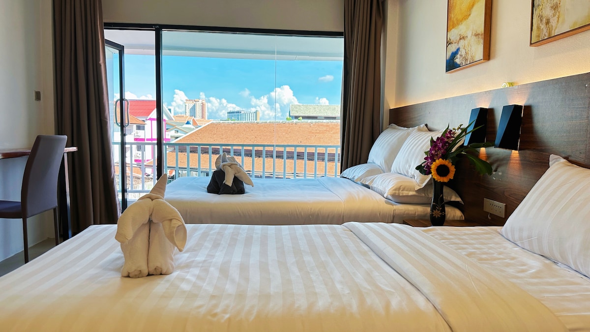 A twin bedroom features two spacious beds with crisp linens, complemented by decorative towel art shaped like elephants. A large sliding door opens to a balcony, revealing a view of the city skyline under a bright blue sky. Natural light fills the room.