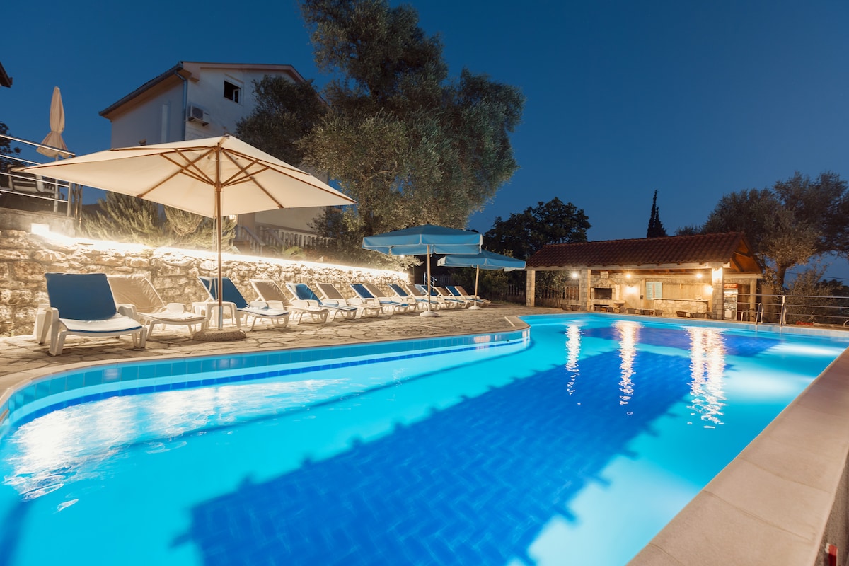 A swimming pool is illuminated at night, surrounded by deck chairs and umbrellas. Soft lighting highlights the surrounding landscaping, including olive trees. A separate building with a gabled roof is visible in the background, enhancing the serene atmosphere.