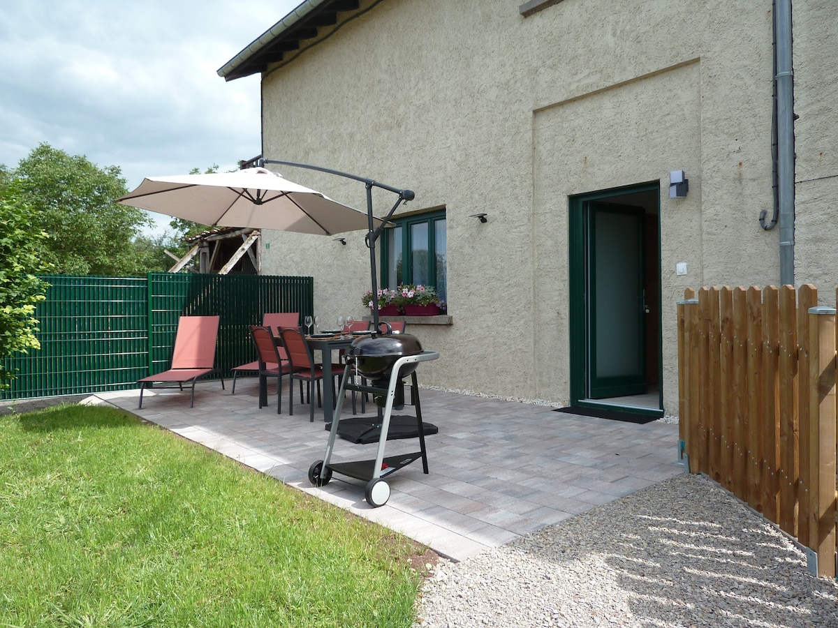 An outdoor terrace is presented with a dining table and four chairs, complemented by a large umbrella for shade. A grill is positioned nearby, and a fence defines the space. Lush greenery surrounds the area, enhancing the outdoor experience.