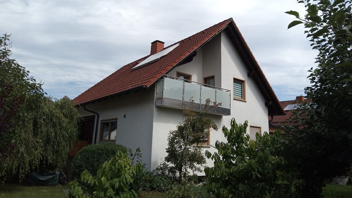 The exterior of the property is depicted, showcasing a two-story building with a sloped roof and a balcony featuring a glass railing. Surrounding greenery includes well-maintained shrubs and trees, providing a natural setting. A clear sky with some clouds is present in the background.