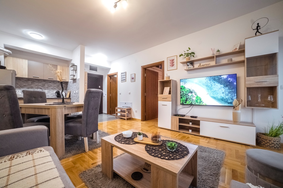 A modern living space features a comfortable seating area with gray chairs and a spacious coffee table. A large television is mounted on a light wood entertainment unit. The dining area, visible in the background, showcases a round table surrounded by four chairs.