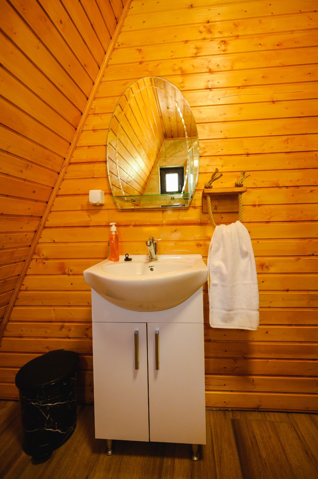 A compact bathroom features a white sink mounted on a sleek cabinet, complemented by a round mirror above. A towel hangs on a hook to the side, while a small black trash bin is positioned beneath the sink. Warm wooden walls add a natural touch.