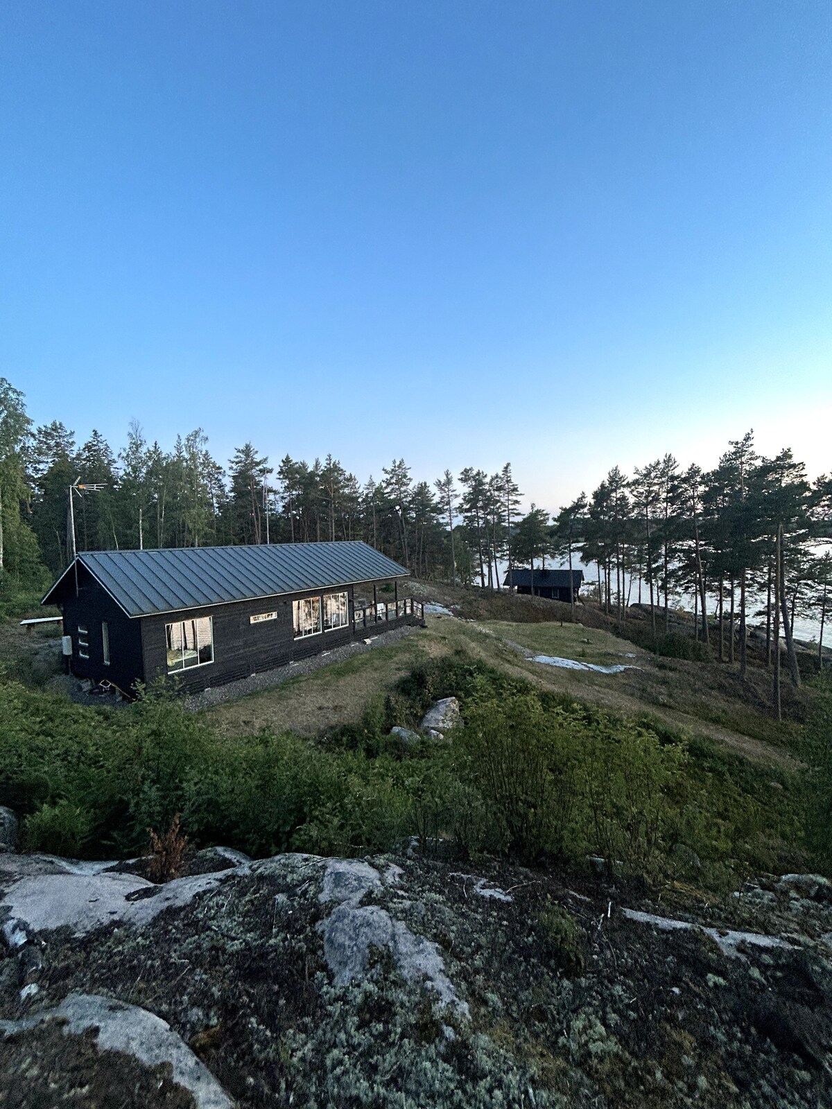 Two cabins are nestled among trees on a hillside, overlooking the water. One cabin has a black exterior with large windows, while the other is more distant, partially obscured by foliage. The natural landscape includes grassy areas and rocky outcrops.