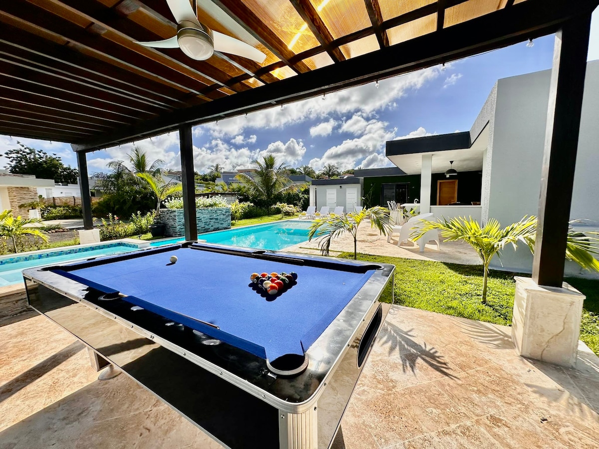 An outdoor living area showcases a sleek pool table positioned under a covered terrace. A shimmering private pool is visible in the background, surrounded by lush greenery and palm trees, enhancing the relaxing atmosphere of the space.