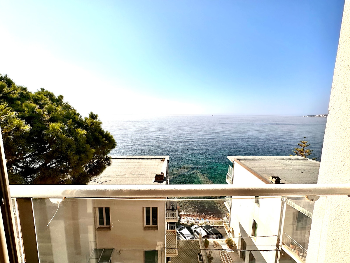 A clear view of the tranquil sea extends from the balcony, showcasing the horizon where the water meets the sky. The sunlight reflects off the surface, creating a serene ambiance. Lush greenery is visible from the side, framing the scene.