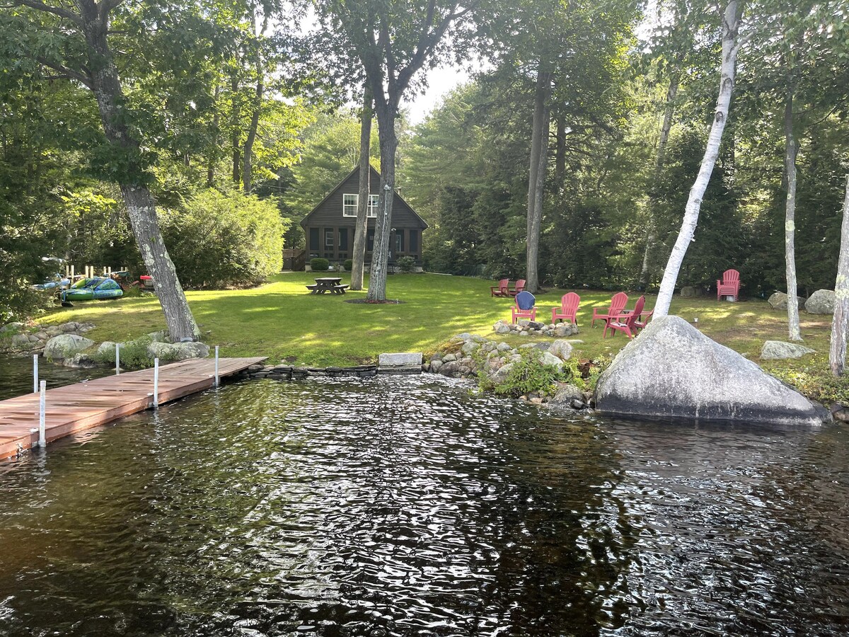 A serene lakeside view highlights a charming cottage surrounded by lush trees. A grassy area features several red Adirondack chairs and a picnic table. The wooden dock extends into the water, creating a peaceful outdoor space to enjoy nature.