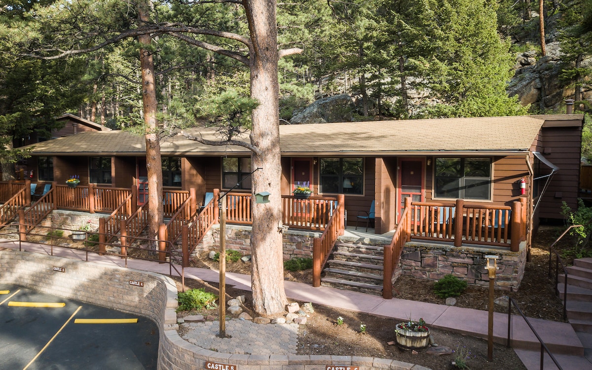 A cluster of cozy motel rooms is set against a backdrop of tall trees and rocky terrain. Each room features a covered front patio with wooden railings, and stone steps lead to the entrance. A paved driveway is visible in the foreground.