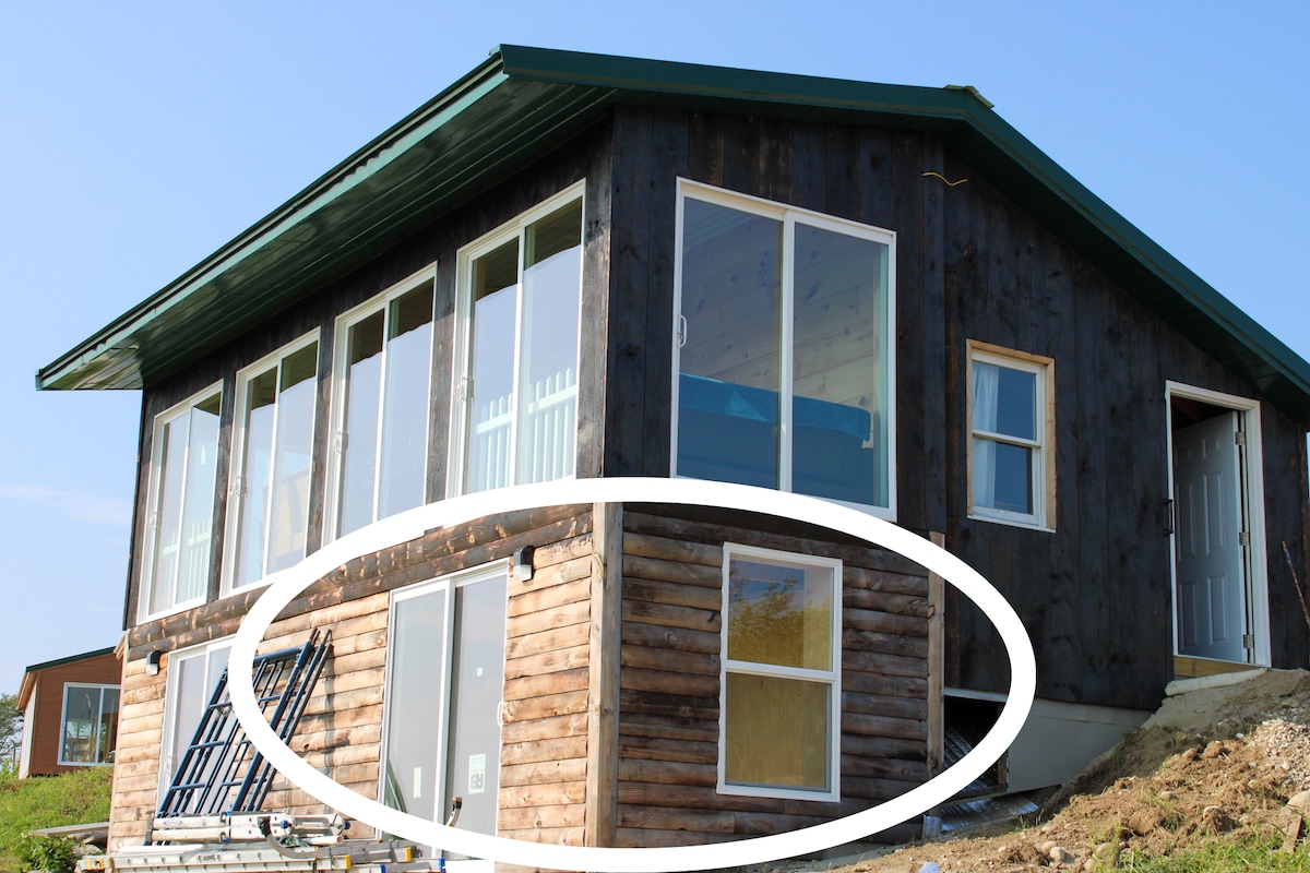 The exterior of the cabin is shown, featuring a combination of wood and large glass windows. The structure is elevated slightly above ground, with a door on the right and a series of windows inviting natural light. A light blue bed frame is visible through the glass.