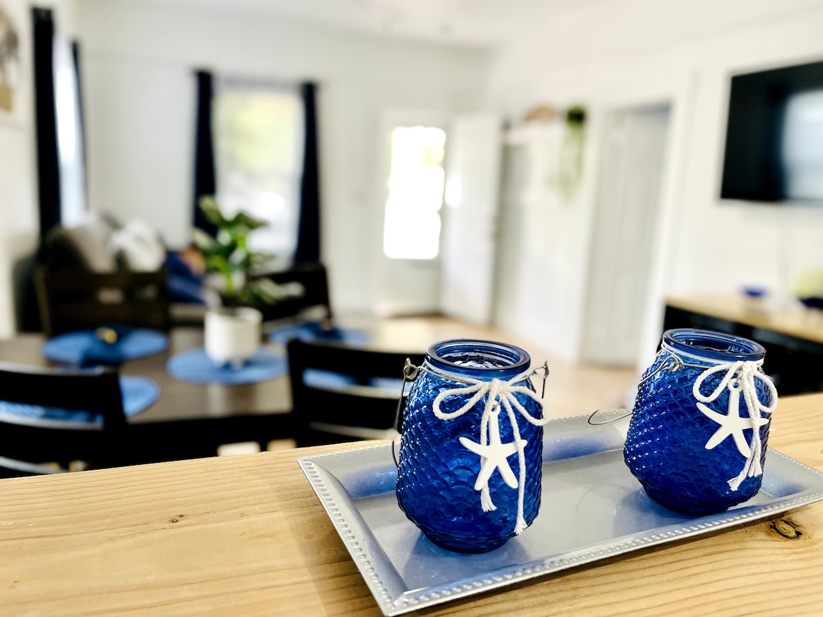 Two blue glass candle holders with decorative starfish sit on a silver tray, positioned in the foreground. In the background, a dining area with a round table and black chairs is visible, along with a cozy living space featuring a television and plants.