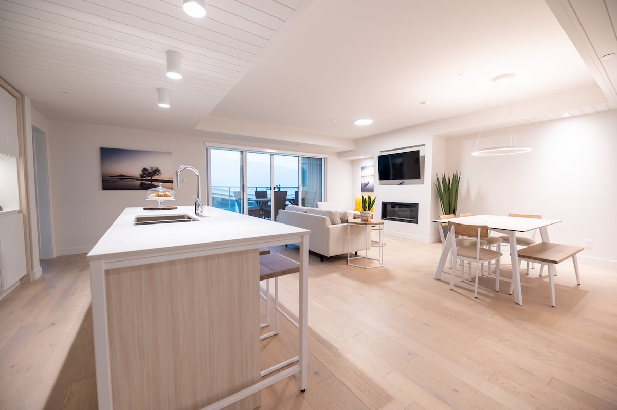 A spacious interior is showcased, featuring a modern kitchen island with seating. A comfortable seating area is visible alongside a dining table. Natural light floods the room through a large sliding door, highlighting neutral tones and sleek furnishings.