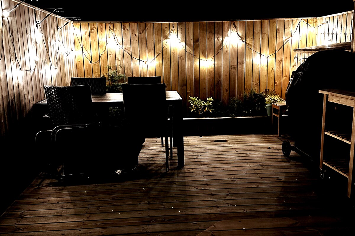 A private patio is illuminated with soft lights, creating a cozy atmosphere. A dining table is surrounded by four chairs. A barbecue grill and wooden storage cart are also present, alongside a planter featuring greenery. The wooden floor adds warmth to the space.