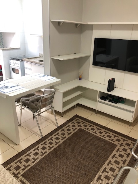 A modern living space features a small dining area with a white table and two silver chairs positioned beside a cozy rug. A wall-mounted flat-screen television is displayed on an entertainment unit, while a glimpse of the kitchen is visible in the background.