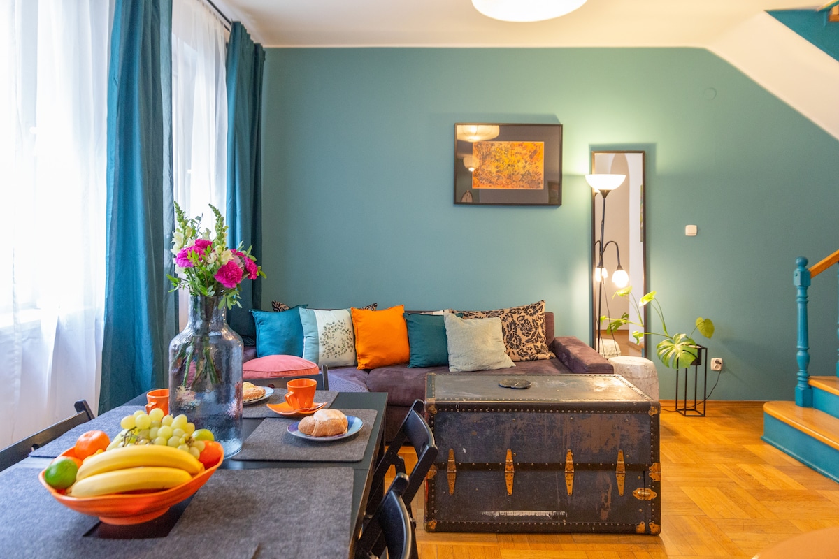 A bright living room is depicted, featuring a cozy lounge with a dark sofa adorned with colorful cushions. A dining table is set with fresh fruit, pastries, and vibrant tableware. Light floods in through large windows, enhancing the space's inviting atmosphere.