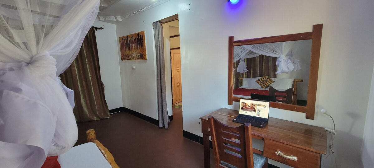A guest room is presented with a wooden desk and chair positioned near a mirror. Soft light from a lamp illuminates the space. A bed with a canopy is visible in the background, alongside decorative pillows. Rich curtains frame the window, enhancing the room's comfort.